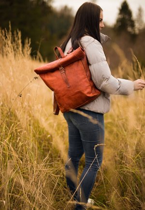 Brown leather backpack with a roll-top design, adjustable straps, and metal hardware, worn by a person in a gray puffer jacket and jeans.