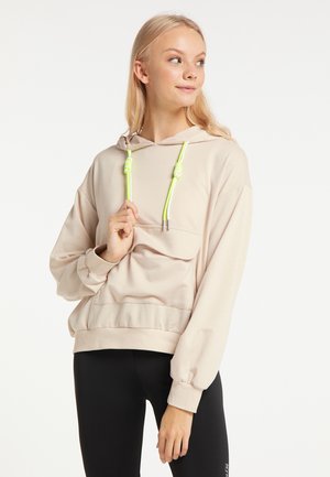 Femme blonde portant un sweat à capuche beige avec des cordons jaunes néon et des leggings noirs, regardant sur le côté en tenant un cordon à la main.