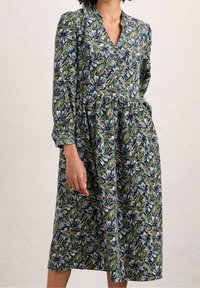 Květinové šaty s dlouhými rukávy, výstřihem do V a nařaseným pasem. Obsahují modré, zelené a bílé květinové vzory na tmavém pozadí. Měkká látka.