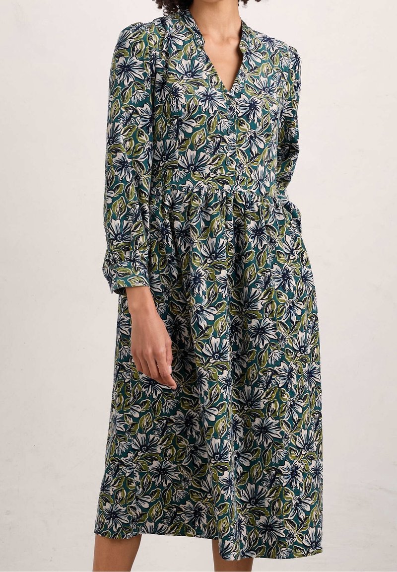 Květinové šaty s dlouhými rukávy, výstřihem do V a nařaseným pasem. Obsahují modré, zelené a bílé květinové vzory na tmavém pozadí. Měkká látka.