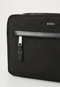 Juodas audinys nešiojamojo kompiuterio krepšys su lygiu paviršiumi, pasižymintis sidabriniu užtrauktuku, juodu oda akcentu ir prekės ženklu "BOSS" išdegtu baltai.