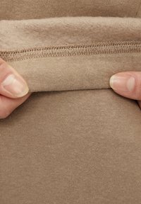Béžová tkanina s měkkou strukturou, která ukazuje složený okraj odhalující huňatou podšívku a prošité švy. Materiál vypadá pružně a pohodlně.