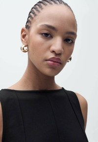 Top sans manches noir au design épuré et minimaliste. Le mannequin porte de grandes boucles d'oreilles en or et a des cheveux tressés en cornrows.