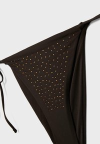 Bas de bikini triangulaire noir avec liens sur les côtés et petits clous dorés disposés en motif diagonal sur le tissu.