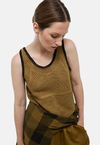 Débardeur en tricot couleur brun clair avec un motif texturé et des bordures noires, associé à un foulard à carreaux noué en vert foncé et noir.