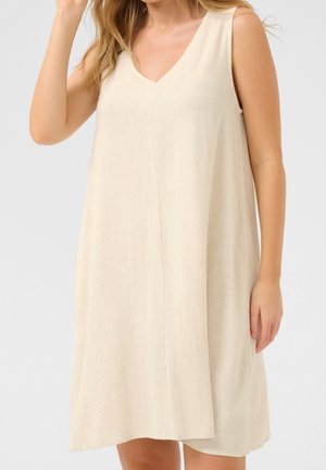 Femme portant une robe beige sans manches, longueur genou, forme évasée, avec un décolleté en V, debout devant un fond gris clair uni.