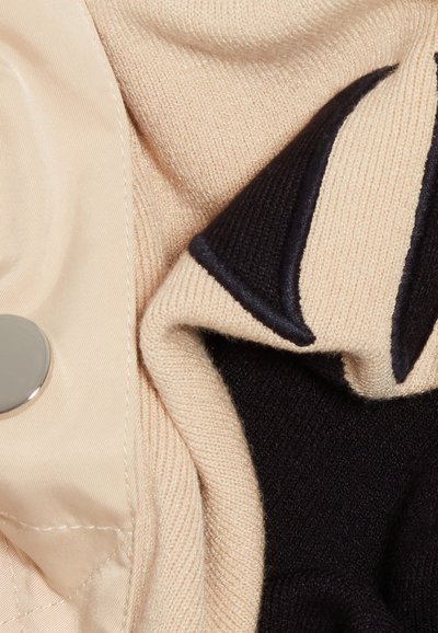 Pull en tricot en tissu beige et noir, texture lisse, design plié, et un motif contrastant avec un accent de bouton métallique.