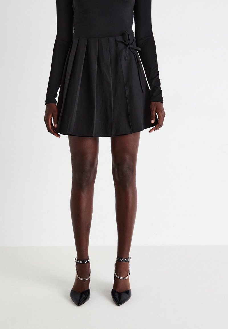 Femme portant une mini-jupe plissée noire avec un nœud sur le côté, un haut noir à manches longues et des talons pointus noirs avec des brides à la cheville et des chaînes.