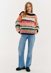 Wielokolorowy sweter z poziomymi paskami w pastelowymi i ciemnymi tonacjach, zestawiony z jasnoniebieskimi spodniami dzwonami i czarnymi butami.