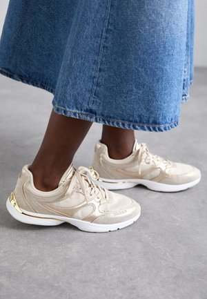 Sneakers atletici beige con dettagli in rete e metallici, punta arrotondata e suole bianche, abbinati a una gonna di jeans blu.