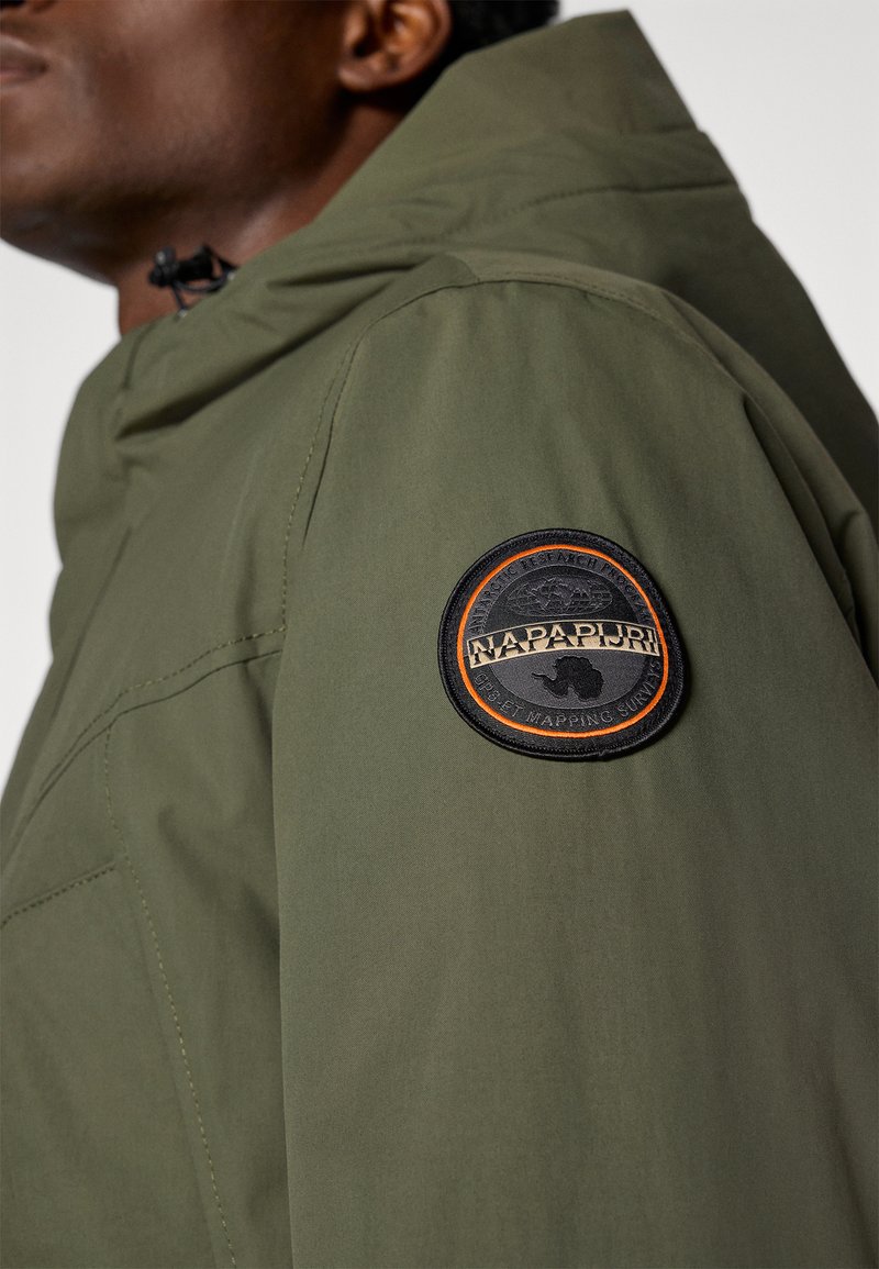 Veste vert olive avec une capuche et un patch rond noir et orange sur l'épaule. Le tissu semble lisse avec des détails de couture visibles.