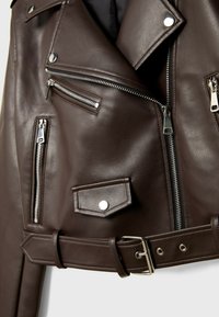 Giacca di pelle marrone scuro con numerosi zip argentati, tasca con bottone a pressione e cintura coordinata con fibbia in metallo.