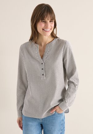Vrouw die glimlacht en een lange mouw draagt, een zwart-wit verticaal gestreept overhemd met knopen en lichtblauwe spijkerbroek, staand tegen een effen achtergrond.