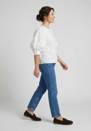 Frau geht nach rechts, trägt eine weiße Bluse mit Puffärmeln, blaue Jeans und dunkelbraune Loafers vor hellem, einfachem Hintergrund.