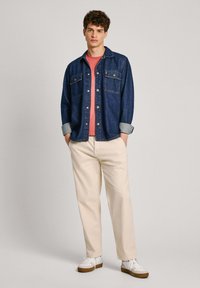 Giacca di jeans con due tasche sul petto, maniche arrotolate, indossata sopra una camicia rossa. Pantaloni beige chiaro e sneakers bianche con accenti marroni.