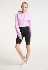 Lila crop hoodie med elastiskt midjeband, långa ärmar och huva, parat med svarta cykelshorts med logodetaljer. Vita sneakers.
