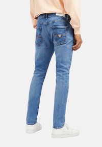 Jeans en denim bleu clair avec une coupe slim, comportant deux poches arrière et un détail de logo sur la taille. Portés avec des baskets blanches.