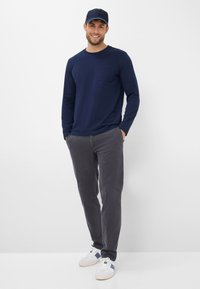 Navy langemouwen shirt met een zak, grijze broek en witte sneakers met blauwe accenten. Man staat zelfverzekerd voor een witte achtergrond.