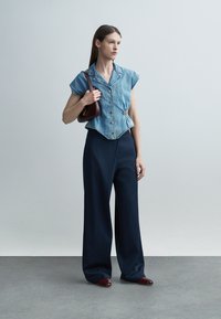 Femme portant un haut bleu en denim à manches courtes avec boutons, un pantalon large bleu marine, des chaussures bordeaux et un petit sac à bandoulière marron.