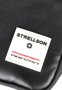 Svart tygpåse med en vit etikett som visar "STRELLSON", en logotyp och koordinater. Har en texturerad yta och sydda detaljer.