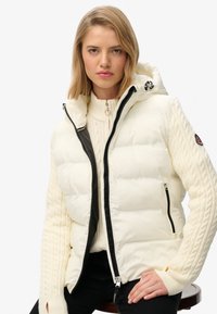 Biały kamizelka puchowa z kapturem i czarnym zamkiem błyskawicznym. Posiada rękawy z plecionym wzorem w warkocz oraz dwie boczne kieszenie.