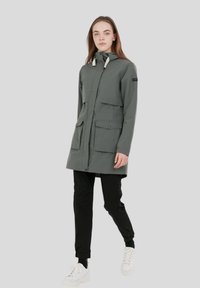 Parka lunga grigio-verde con cappuccio, due tasche frontali, chiusura a pressione frontale e cordini regolabili. Il modello indossa pantaloni neri e sneakers bianche.