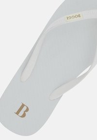 Baltā flip-flop čība ar teksturētu pelēku zoli. Plānas baltas siksnas ar zelta logo "B" uz virsmas. Minimālisma dizains.