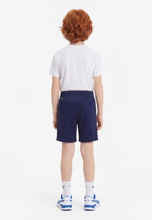 Barn med rødt krøllet hår, der står med fronten væk, iført en hvid t-shirt, navyblå shorts, hvide Puma-strømper og blå og hvide sneakers.