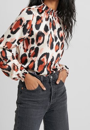 Femme portant un chemisier à manches longues et col montant avec un imprimé léopard rouge, noir et beige, rentré dans un jean taille haute gris foncé.