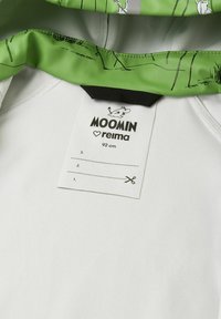 Biała odzież z zielonym kołnierzykiem z rysunkami Muminków. Metka zawiera nazwę marki, rozmiar (92 cm) oraz instrukcje dotyczące kroju.