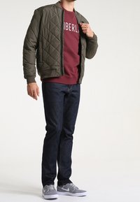 Giubbotto bomber imbottito verde oliva su una felpa bordeaux con scritte bianche, abbinato a jeans scuri e sneakers grigie.