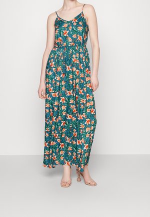 Kvinde iført ærmeløs teal maxikjole med orange og hvide blomstermønstre samt beige åbne hælede sandaler, stående foran en ensfarvet hvid baggrund.