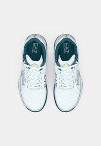 Fehér sportcipő texturált mintával, türkizkék díszítésekkel és logóval a nyelvén. Kerek fűzőlyukak és párnázott talp jellemzi.