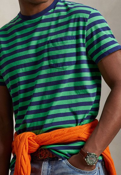 T-shirt rayé vert et marine avec une poche, associé à un pull orange en maille torsadée noué à la taille. Jean en denim et montre marron complètent le look.