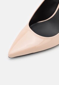 Chaussure à talon haut en cuir verni nude à bout pointu avec doublure intérieure noire sur fond blanc.