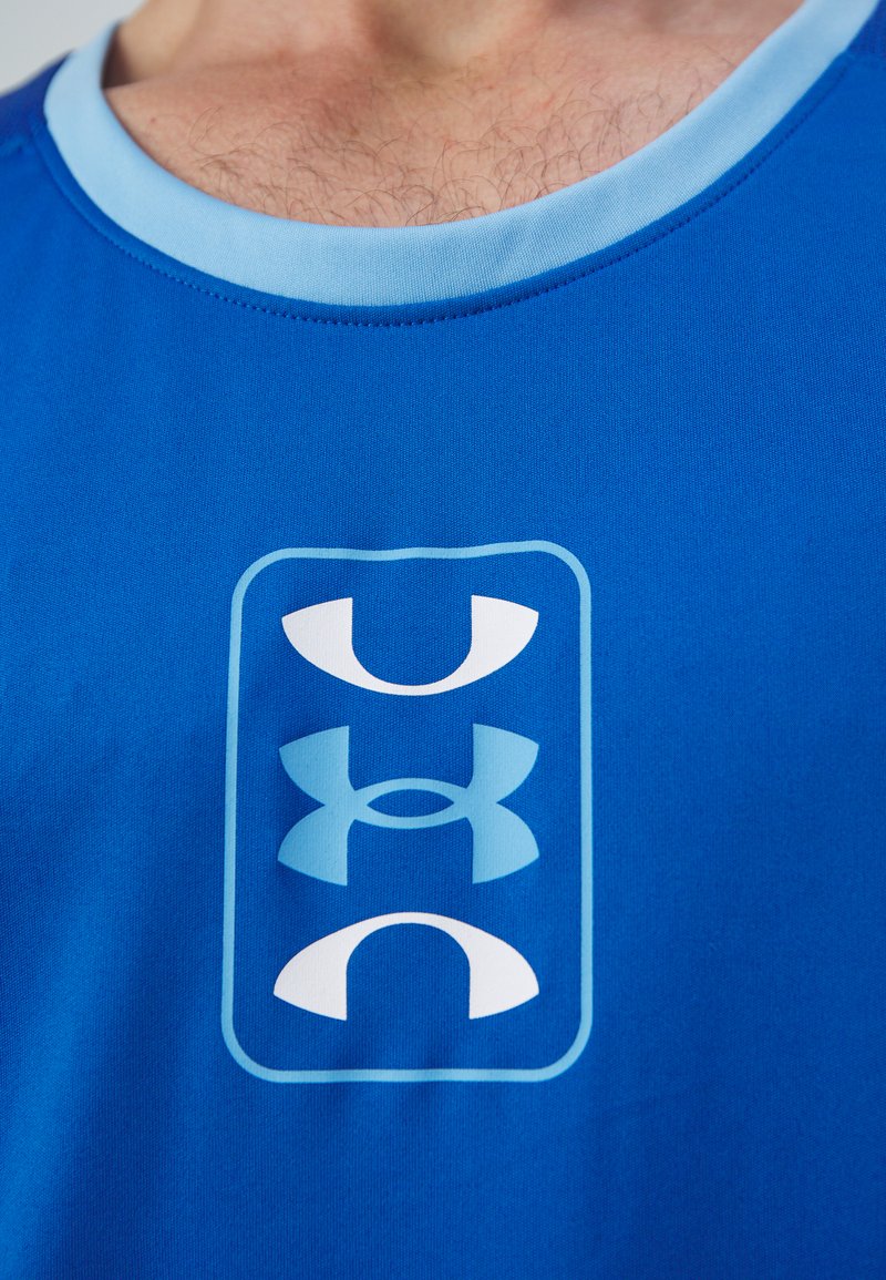Niebieski sportowy top na ramiączkach z jasnoniebieską kołnierzykiem. Z przodu znajduje się duża biała grafika z nałożonymi na siebie logo. Gładka tekstura materiału.
