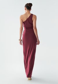 Bordové šaty na jedno rameno z hladkej látky, priliehavé v páse, s dlhou sukňou až po zem a elegantným bočným detailom. Zlatý náramok ako doplnok.