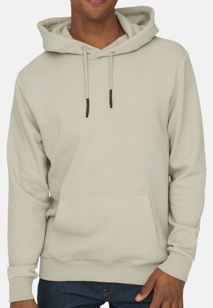Mand iført lysebeige pullover-hoodie med frontlomme og snore, kombineret med mørkeblå jeans, synlig fra hagen til hofterne.