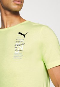 Camiseta amarilla de manga corta con un logo negro de Puma, con texto impreso en azul. El material parece suave y ligero.