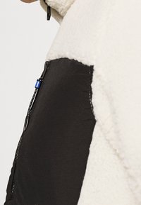 Giacca fleece con pannelli in nylon neri, colletto alto e chiusura con zip dotata di un tassello blu. Tessuto morbido e testurizzato.