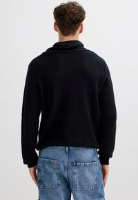 Suéter negro de punto con cuello alto, combinado con pantalones vaqueros azul claro que tienen grandes bolsillos traseros. La textura es acanalada y ligeramente voluminosa.