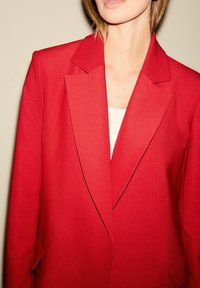 Rød blazer med et struktureret design, skarpe revers og let glans. Lavet af glat stof, med enkeltknap lukning og klaftlommer.