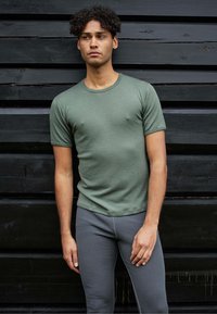 Grøn kortærmet t-shirt i et blødt, tekstureret stof parret med grå tætsiddende bukser, sat op mod en sort træbaggrund.