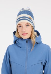 Strickmütze mit blauen und elfenbeinfarbenen Streifen, mit einem Etikett. Kombiniert mit einer blauen Softshelljacke, mit Kapuze und mit Reißverschluss versehenem Brusttasche.