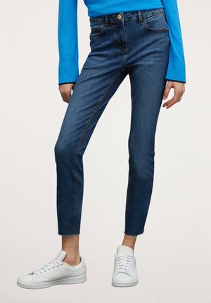 Personne portant un jean slim bleu foncé avec des ourlets bruts, un haut à manches longues bleu vif, et des baskets blanches, debout devant un fond uni.