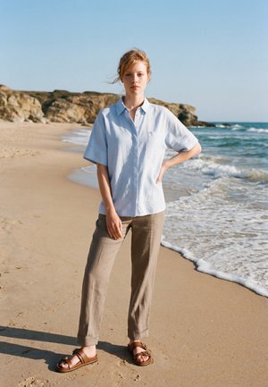 Femme debout sur une plage de sable près du bord de l'eau, portant une chemise bleu clair à manches courtes, un pantalon beige et des sandales marron.