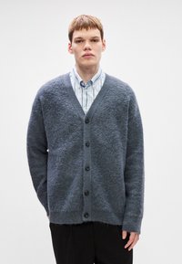Gray wool cardigan with a v-neck, front buttons, and long sleeves. Soft texture with a slightly frayed hem. Worn over a blue striped shirt.