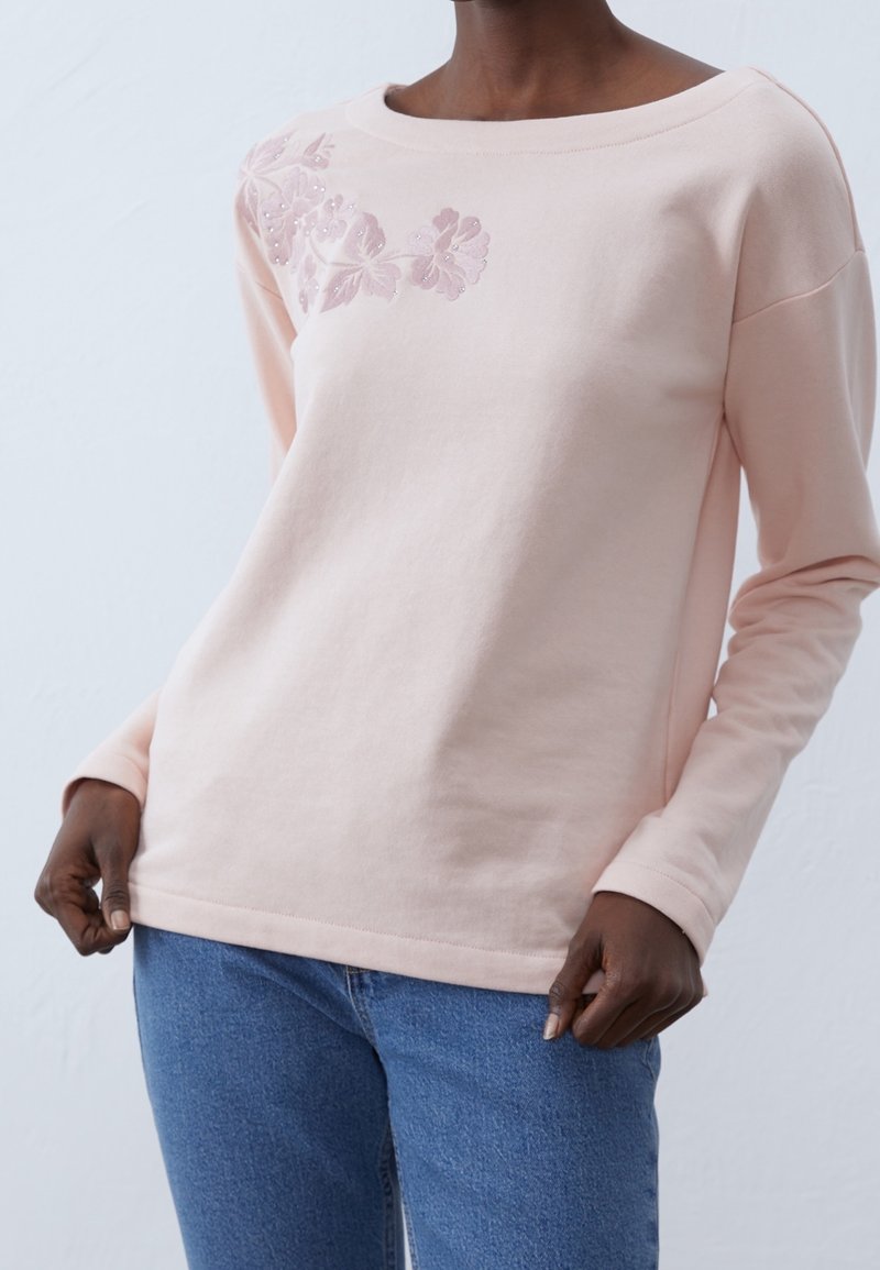 Personne à la peau foncée portant un haut à manches longues rose clair avec broderie florale sur l'épaule et un jean bleu, debout devant un fond uni.
