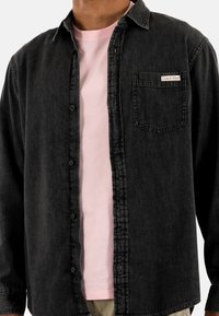 Camisa de mezclilla negra con cierre de botones, cuello tipo polo y bolsillo en el pecho, superpuesta sobre una camiseta de color rosa claro. Acabado casual y con textura.
