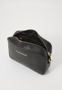 Trousse de maquillage en cuir noir avec une fermeture éclair et des accessoires en or, de forme arrondie et portant le mot "VALENTINO" en relief à l'avant.